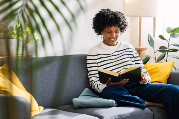 Happy beautiful African American woman reading a book sitting on sofa at home on a cozy winter afternoon